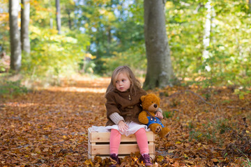 Adorable little girl in the forest with her teddy bear sitting on a wooden chest