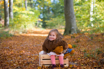 Adorable little girl in the forest with her teddy bear sitting on a wooden chest