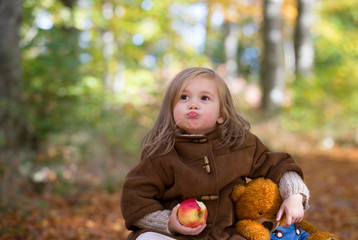Fairy little girl sitting on a wooden chest in the forest with her teddy bear and eating on apple,funny face expressions