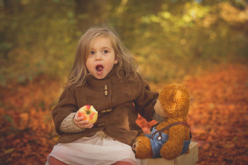 Fairy little girl sitting on a wooden chest in the forest with her teddy bear and eating on apple,funny face expressions