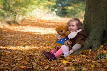 Fairy little girl in the autumn forest sitting near tree with her teddy bear