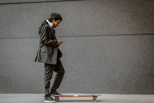 Young urban dreadlocks hipster man standing with one foot on his long board and holding his telephone. Standing on street with black wall in background.