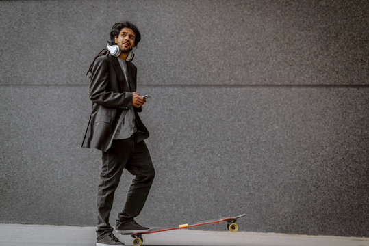Young urban dreadlocks hipster man standing with one foot on his long board and holding his telephone. Standing on street with black wall in background.