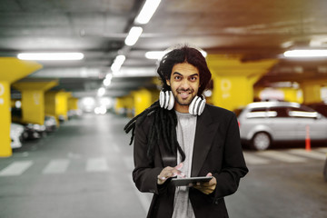 Young cute urban hipster man holding tablet and looking at camera. Standing in garage with headphones around his neck.