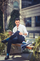 Young stylish businessman uses pc tablet while sitting on bench outside