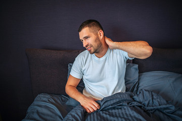 Young man sits on bed and feels pain in neck area. He shrinks and looks to left. Guy holds hand on neck behind. He is partly covered with blanket.