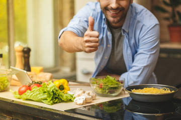 Man preparing delicious and healthy food in the home kitchen on a sunny day. Using tablet computer for searching recipes. Thumbs up.