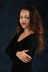 Waist up portrait of a young woman with long curly hair wearing  black  dress with arms crossed on a dark  background in the studio