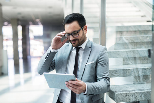 Happy businessman reading news on the tablet device and holding eyeglasses while leaning on the glass. In background stairs.