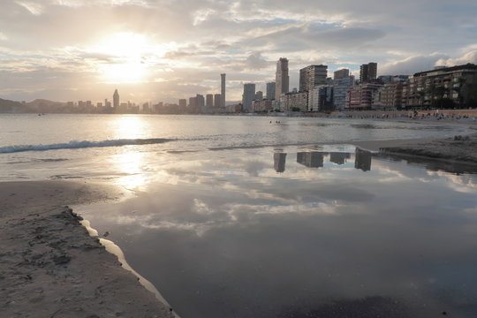Landscape on the shore of the mediterranean sea in Benidorm on the Poniente beach with buildings, the sun at sunset and the clouds reflected in the water