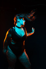 Curvy alternative model with colored hair and lace bodysuit and fishnets poses under blue and orange lighting