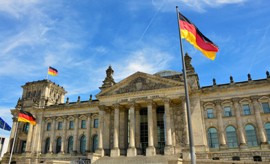 Fototapeta premium Niemieckie flagi powiewające na wietrze w słynnym budynku Reichstagu, siedzibie niemieckiego parlamentu (Deutscher Bundestag), w słoneczny dzień z błękitnym niebem i chmurami, w centralnej dzielnicy Berlin Mitte, Niemcy.