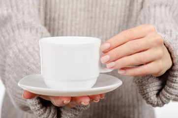 Young woman in sweater holding a white cup and saucer in hands, drinking hot coffee on white background. Space for your logo or text.