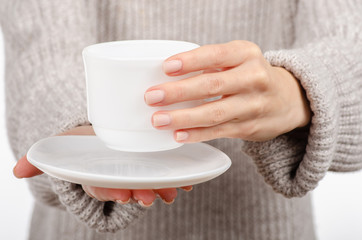 Young woman in sweater holding a white cup and saucer in hands, drinking hot coffee on white background. Space for your logo or text.