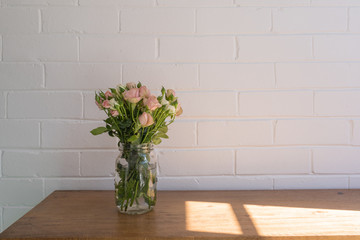 Close up of small pink and cream roses in glass jar on wooden oak sidetable against white painted brick wall with afternoon sunlight