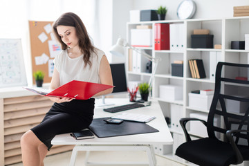 A young girl sat down on a table in the office and holding a red folder.