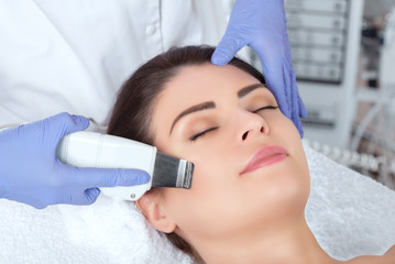 Obraz na płótnie Canvas The doctor-cosmetologist makes the ultrasound cleaning procedure of the facial skin of a beautiful, young woman in a beauty salon. Cosmetology and professional skin care.