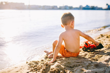 Caucasian child boy playing toy red tractor, excavator on a sandy beach by the river in red shorts at sunset day