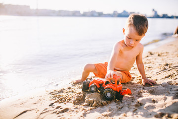 Caucasian child boy playing toy red tractor, excavator on a sandy beach by the river in red shorts at sunset day © Elizaveta