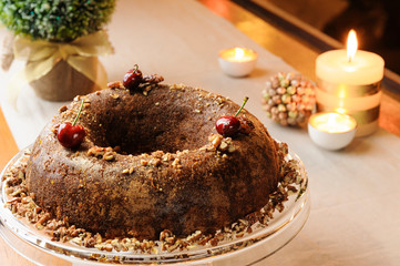 Fototapeta na wymiar Delicious nutty chocolate cake served with cherry on a ceramic dish. Decorated table with candles, small tree and pine cone, on a cloth background