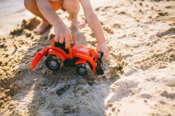 Caucasian child boy playing toy red tractor, excavator on a sandy beach by the river in red shorts at sunset day