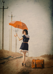 Beautiful brunette girl with suitcase and umbrella on the countryside road.