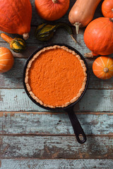 Brightly colored pumpkin pie in cast iron pan surrounded with organic pumpkins and squashes on blue wooden background