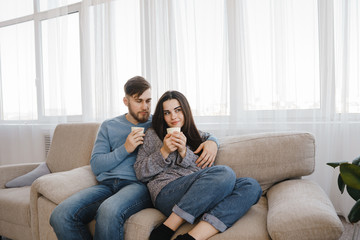 Young couple sitting at sofa with hot drinks. Love, relationships, conversation, winter weekend concept