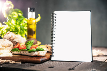 Rustic table with ciabatta bread, avocado, tomatoes, mozzarella, olive oil, fresh basil and notebook with copyspace for your text. Verticall orientation of photo.