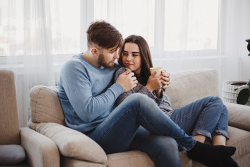 Obraz premium Young couple sitting at sofa with hot drinks. Love, relationships, conversation, winter weekend concept