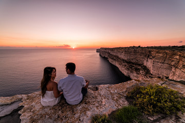 Couple overlooks the famous Ta Cenc Cliffs at sunset.  These cliffs on the island of Gozo overlook the Mediterranean Sea.  Country of Malta. - obrazy, fototapety, plakaty