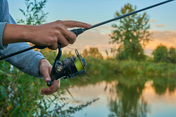 Lake at sunset. A fisherman on the  bank. Fishing rod wheel closeup. Spinning reel. The concept of outdoor activities. Tackles for pike, perch, zander.