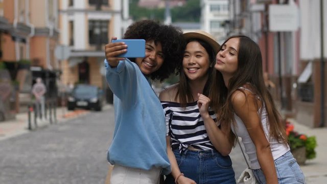 Excited carefree multi ethnic young girlfriends in trendy clothes taking a selfie on smart phone over cityscape background. Cheerful multiracial girls having fun and making self photo on cellphone.
