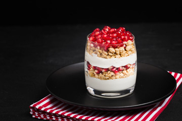 Pomegranate parfait - sweet organic layered dessert with granola flakes, yogurt and fruit seeds on black background.