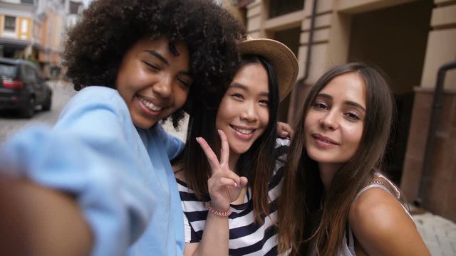 Portrait of beautiful multi ethnic young girlfriends taking a selfie portrait photo on smart phone in city street. Excited diverse hipster girls making funny self portrait on cellphone outdoors.