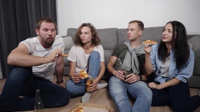 Food, leisure and frienship concept - four smiling young people eating pizza at home and drinking a beer while sitting on the floor. Bearded man telling funny stories, everybody laughing, having good