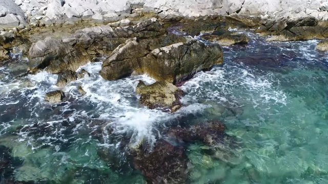 Slow motion aerial view of rocky coastline or seashore is the area where land meets the sea or ocean drone moving left slowly showing the shallow azure blue water and waves splashing at rocks 4k
