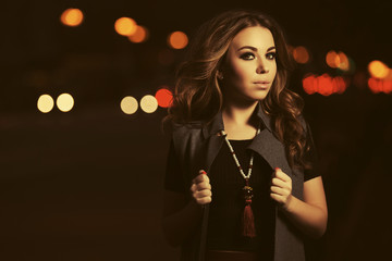 Young fashion woman in long sleeveless coat walking in night city street