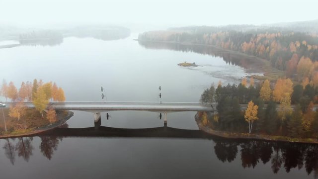 Aerial, tilt down, drone shot, flying towards a car on a bridge, surrounded by mist and autumn colors,, on a foggy, misty and cloudy, fall day, at lake Pielinen, in Nurmes, North Karelia, Finland