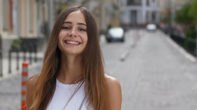 Portrait of gorgeous caucasian girl with long brown straight hair and deep brown eyes smiling at camera outdoors. Joyful young woman with clean tanned skin looking at you with innocent enigmatic smile