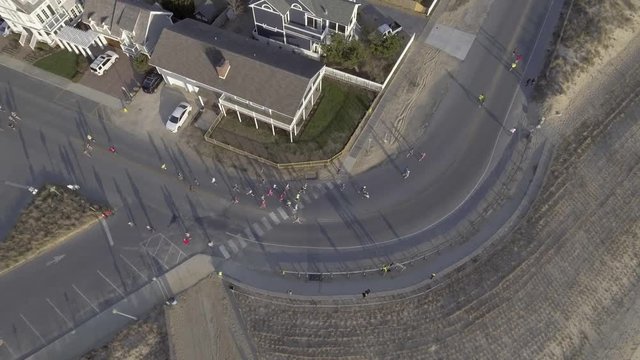Drone/aerial lookdown shot of marathon runners turning corner next to beach as camera rises.