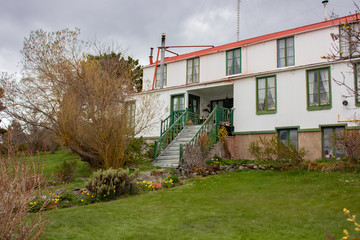 Casa de campo blanca de techo rojo y escaleras con barandas verdes.  Ventanas varias. Flores de colores