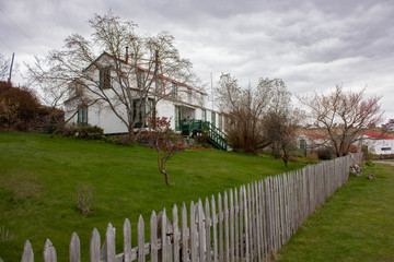 Perspectiva de casa de campo blanca con techo rojo. Escalera principal verde. Césped corto y cerca de madera alineada.
