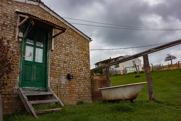 Entrada de casa de campo de ladrillo, con puerta verde. Bañera de blanca oxidada en el frene con casa de fondo. Cielo nublado color gris..