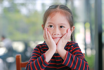 Smiling little Asian child girl with touching her cheek and looking up sitting in the cafe.