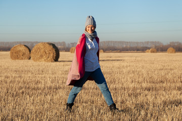 Naklejka na ściany i meble Beautiful modern urban young woman wearing gray knitting hat, pink coat and jeans walking in field, on backgroung haystick. Trendy teenage girl in autumn outdoors.