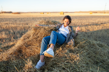 Naklejka na ściany i meble The concept of livestyle outdoor in autumn. Close up of a young woman student in a warm autumn clothes looking funny, smilling, posing, lying on haystick around field