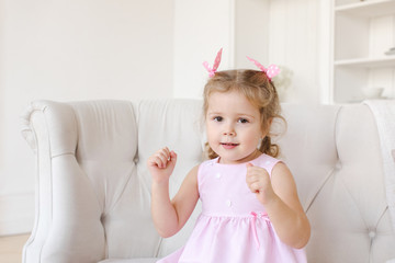 Cute little girl in pink dress looking at camera and gesturing with hands while sitting on comfortable couch in stylish room