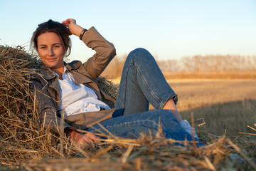 Naklejka na ściany i meble Beautiful modern urban young woman wearing in beige trench, white shirt and jeans lying on a haystack in field. Trendy teenage girl in autumn outdoors.