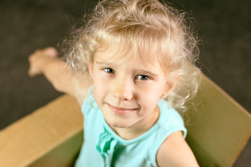 The little blonde smiles inside a cardboard box. shows us hands. dark background. toning. close-up